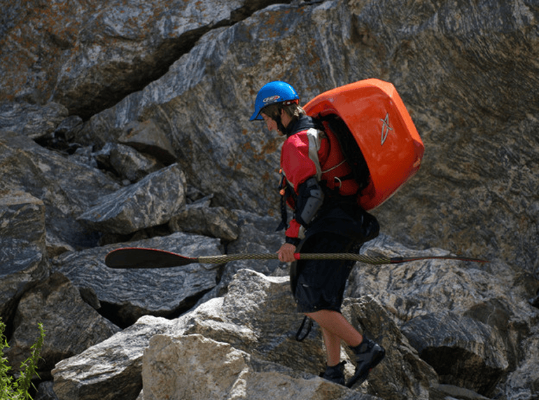 A paddler in a helmet portaging a kayak over rocks beside the river